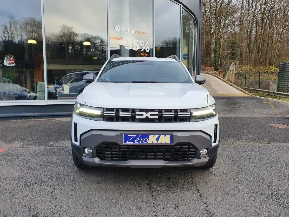 Vue avant d'un Dacia Duster blanc glacier 2025 avec calandre noire et feux LED allumés, stationné devant une vitrine.