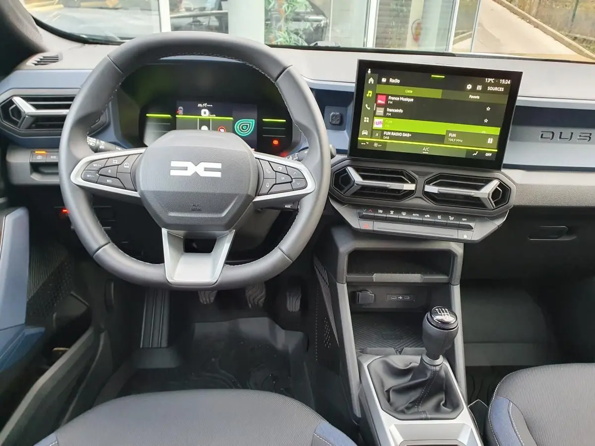 Vue intérieure avant du Dacia Duster 2025 avec volant multifonctions, écran tactile et levier de vitesses manuel.