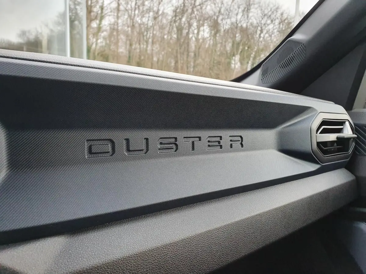 Détail intérieur du tableau de bord noir avec logo DUSTER en relief sur la partie passager du Dacia Duster 2025.