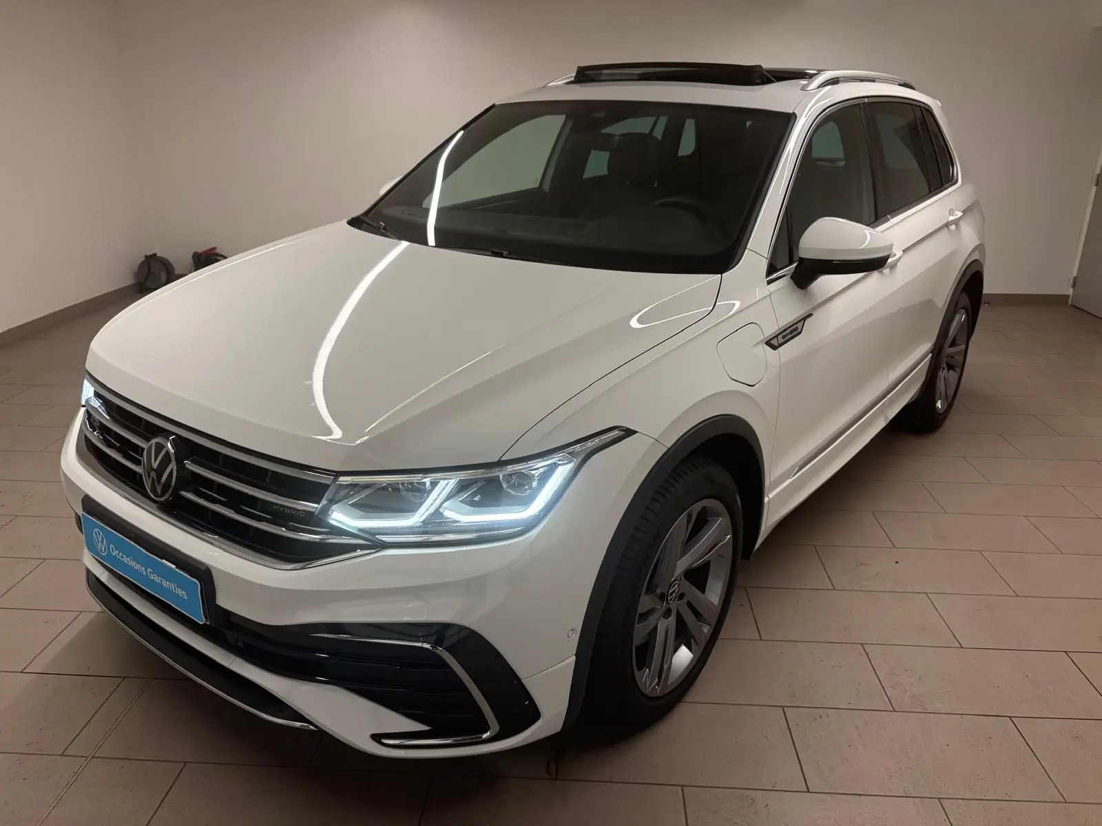 Volkswagen Tiguan blanc pur 2023 en vue 3/4 avant droit avec phares allumés et badge hybride sur aile.