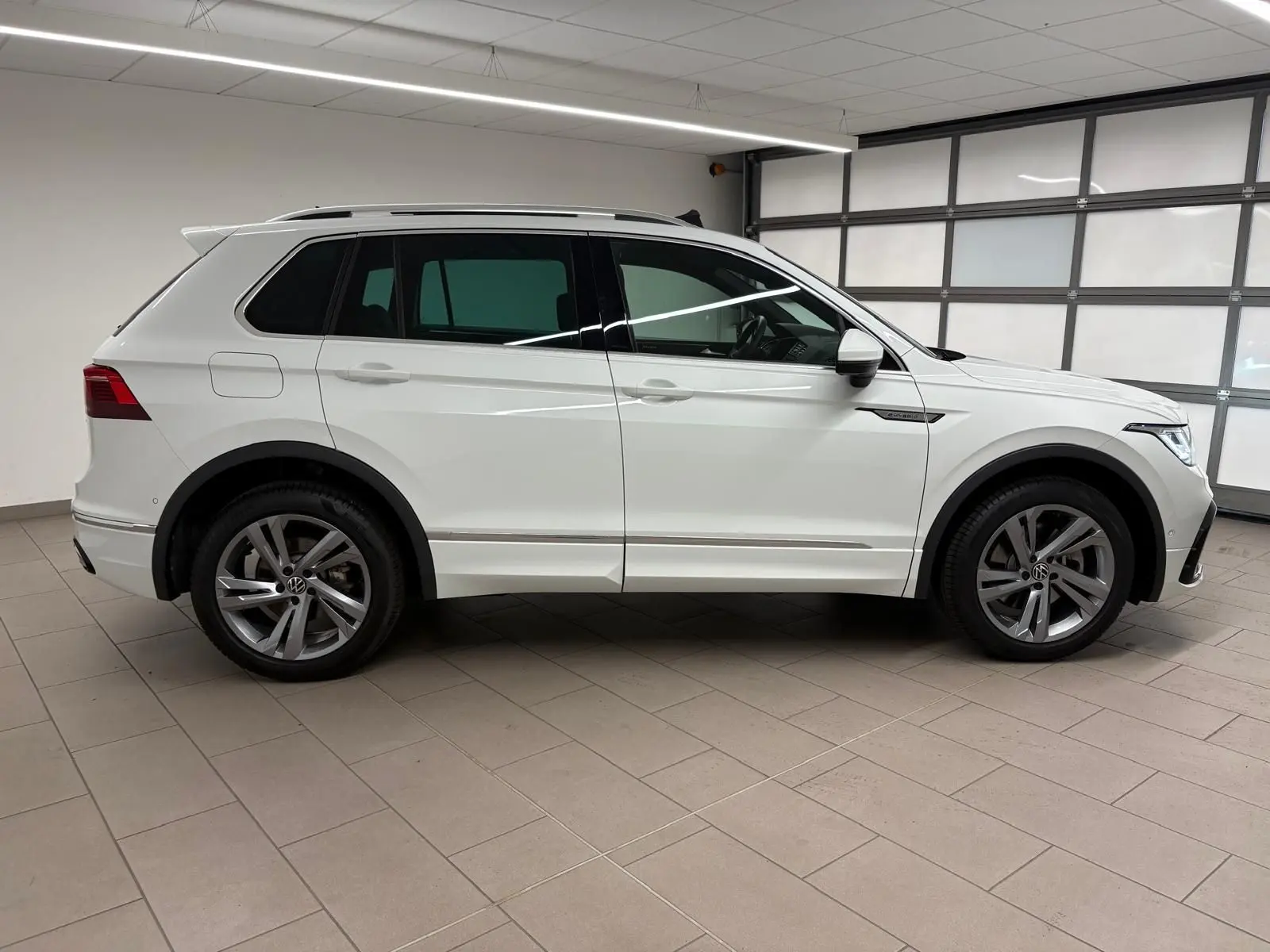 Profil côté gauche d'un Volkswagen Tiguan blanc pur 2023, version hybride rechargeable R-Line Exclusive en intérieur showroom.
