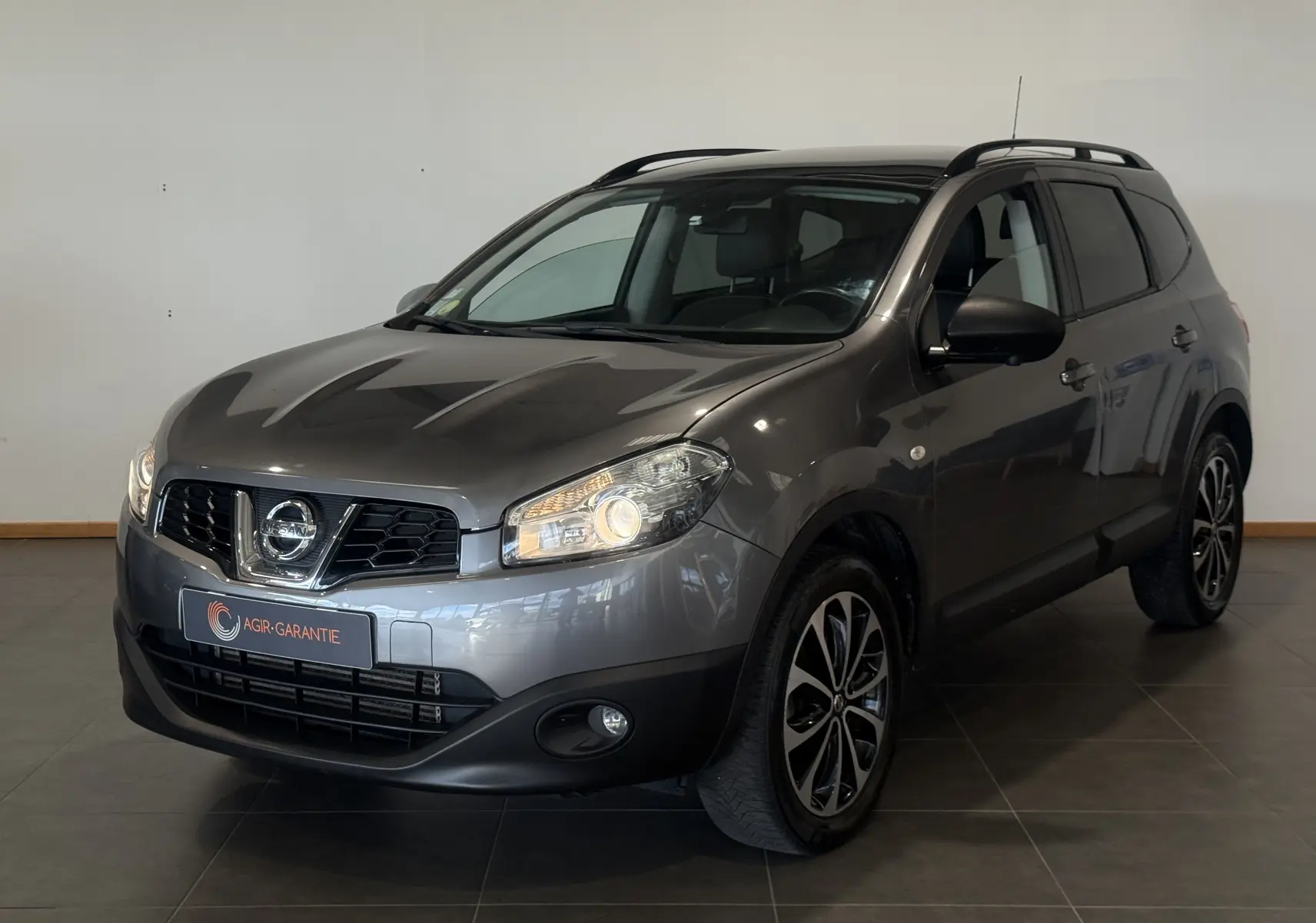 Nissan Qashqai+2 gris 2014 en 3/4 avant droit, avec jantes alliage et phares allumés en intérieur showroom.