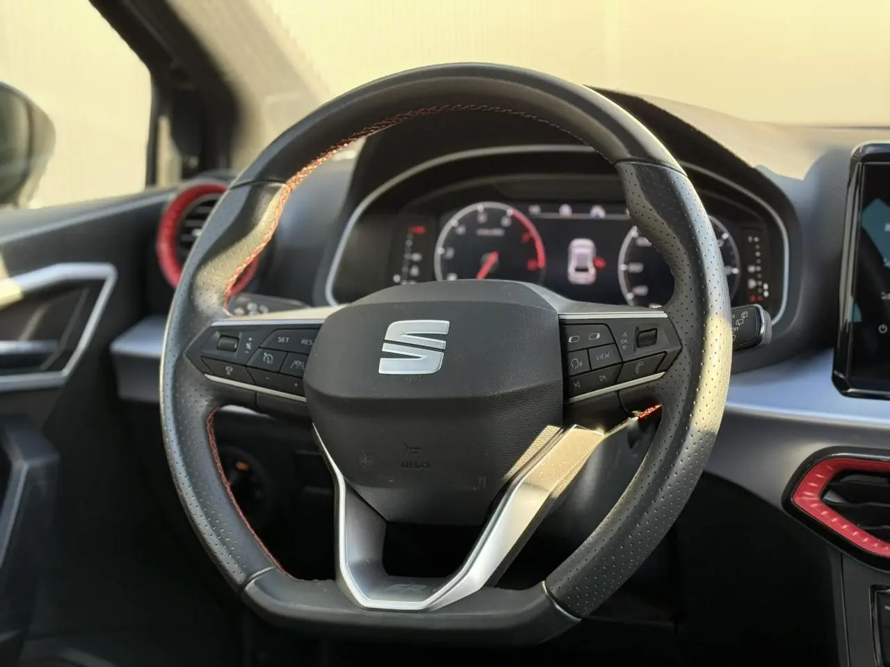 Vue rapprochée du volant perforé noir avec logo SEAT, entouré d'inserts rouges et tableau de bord numérique en arrière-plan.