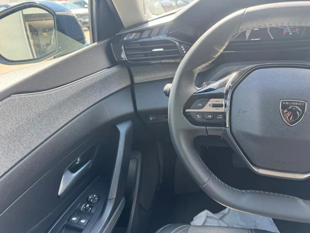 Vue intérieure côté conducteur de la Peugeot 308 Hybrid 136 Allure 2024 noire, volant multifonctions et commandes porte visibles.