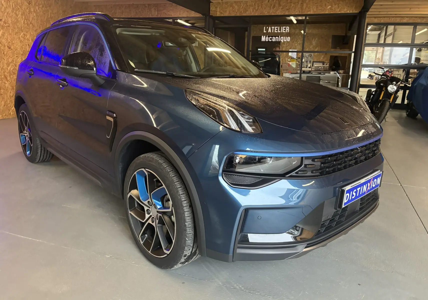 Vue 3/4 avant gauche d'un SUV LYNK & CO 01 bleu avec jantes bicolores et calandre noire en intérieur d'atelier.