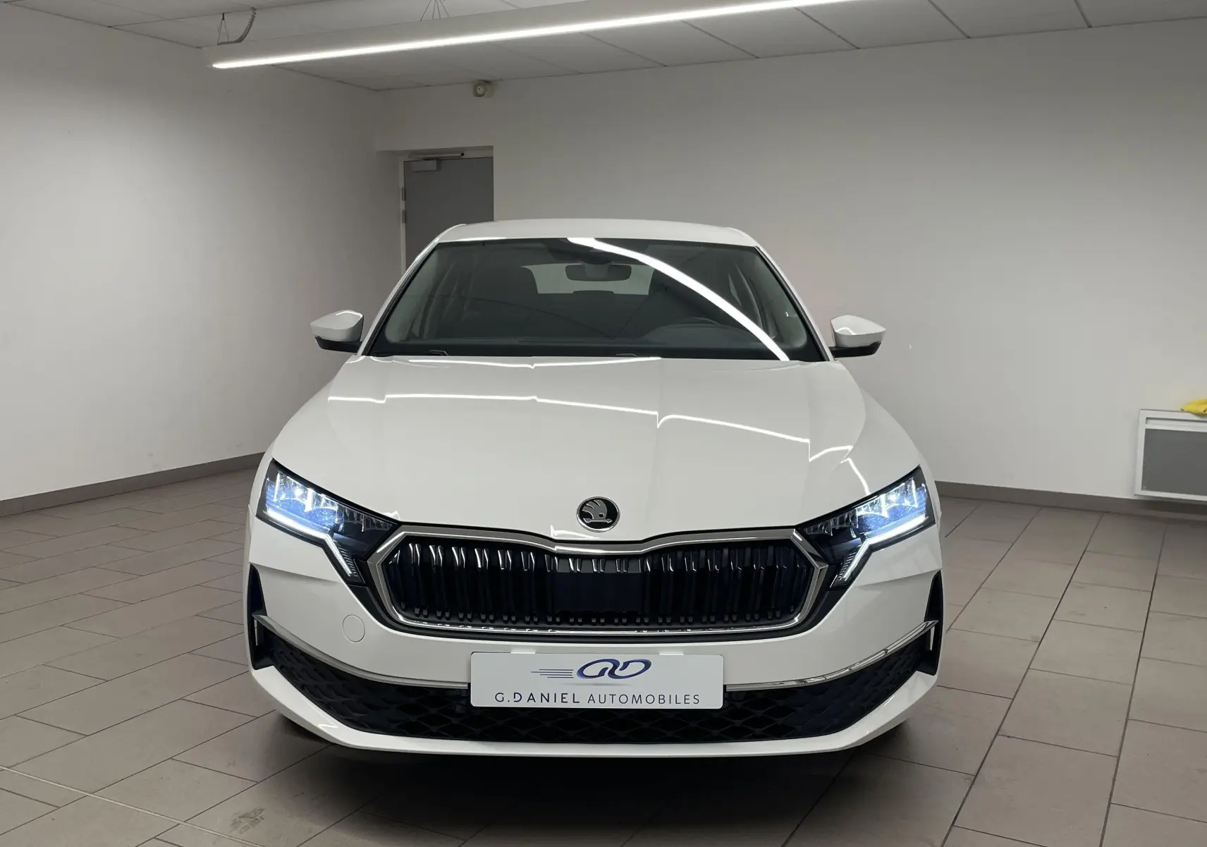 Vue de face d'une Skoda Octavia blanche Candy 2024 avec phares allumés dans un garage intérieur.