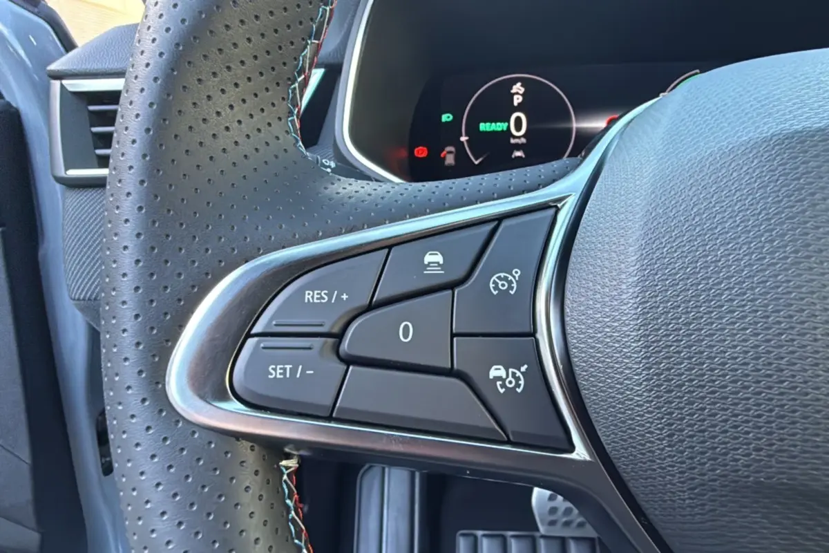 Gros plan sur les commandes au volant perforé de la Renault Clio V E-TECH 145 Esprit Alpine 2025, tableau de bord digital visible.