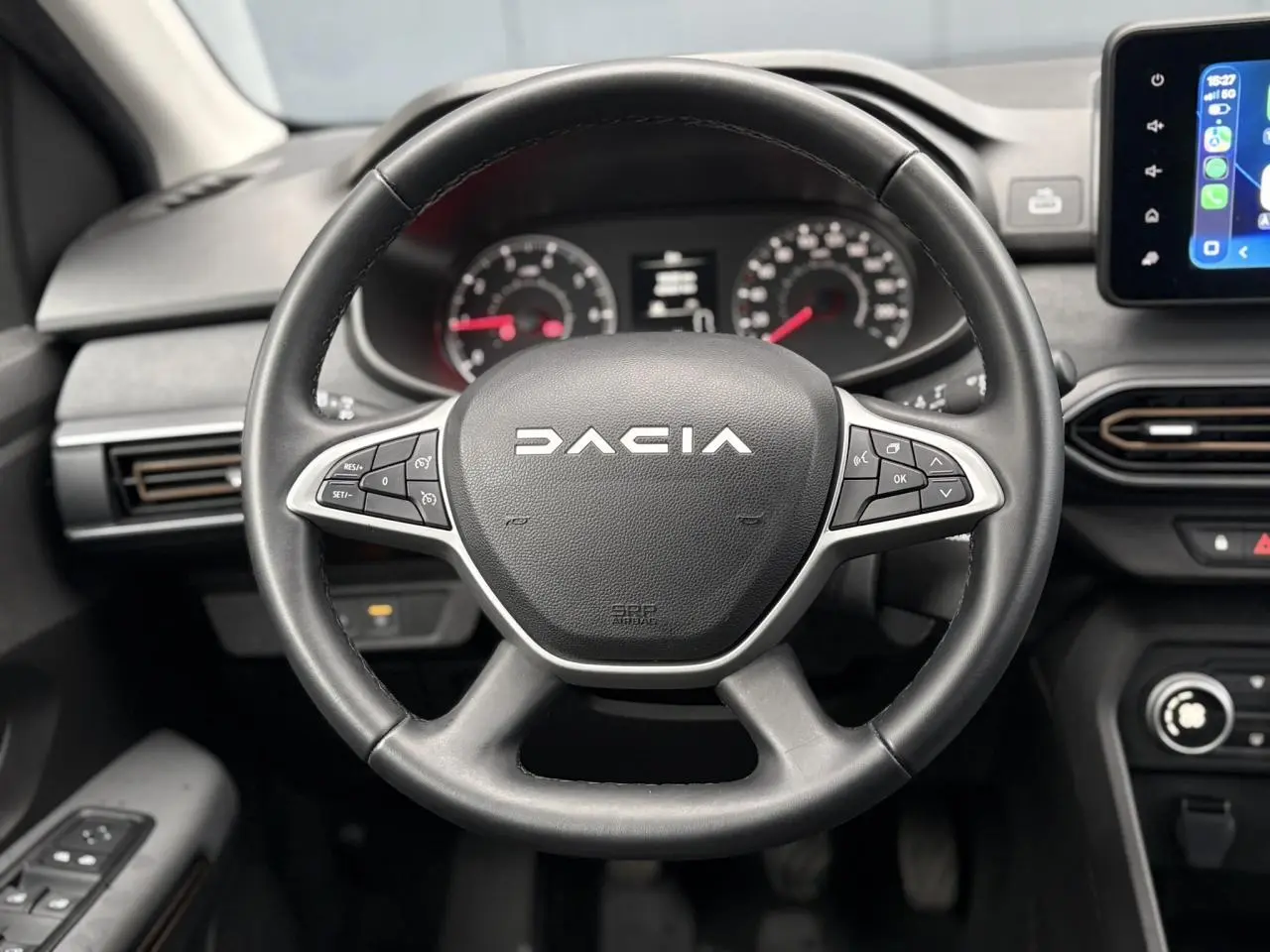 Vue frontale du volant noir Dacia avec commandes intégrées et tableau de bord numérique en arrière-plan.