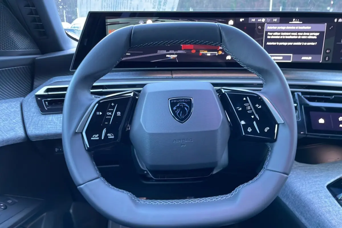 Vue rapprochée du volant cuir multifonctions gris avec logo Peugeot, écran numérique panoramique en arrière-plan.
