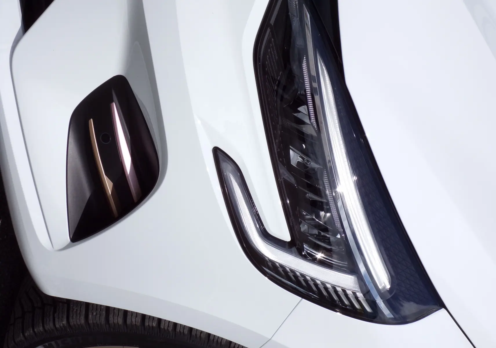 Gros plan sur le phare avant droit et la prise d'air noire avec insert doré d'une Renault Clio V blanche.