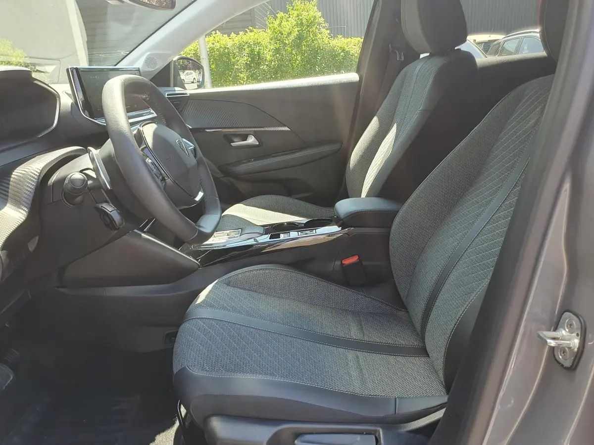 Vue intérieure côté conducteur de la Peugeot 208 1.2 Hybrid 110 Allure gris Sellenium, sièges tissu gris et console centrale brillante.