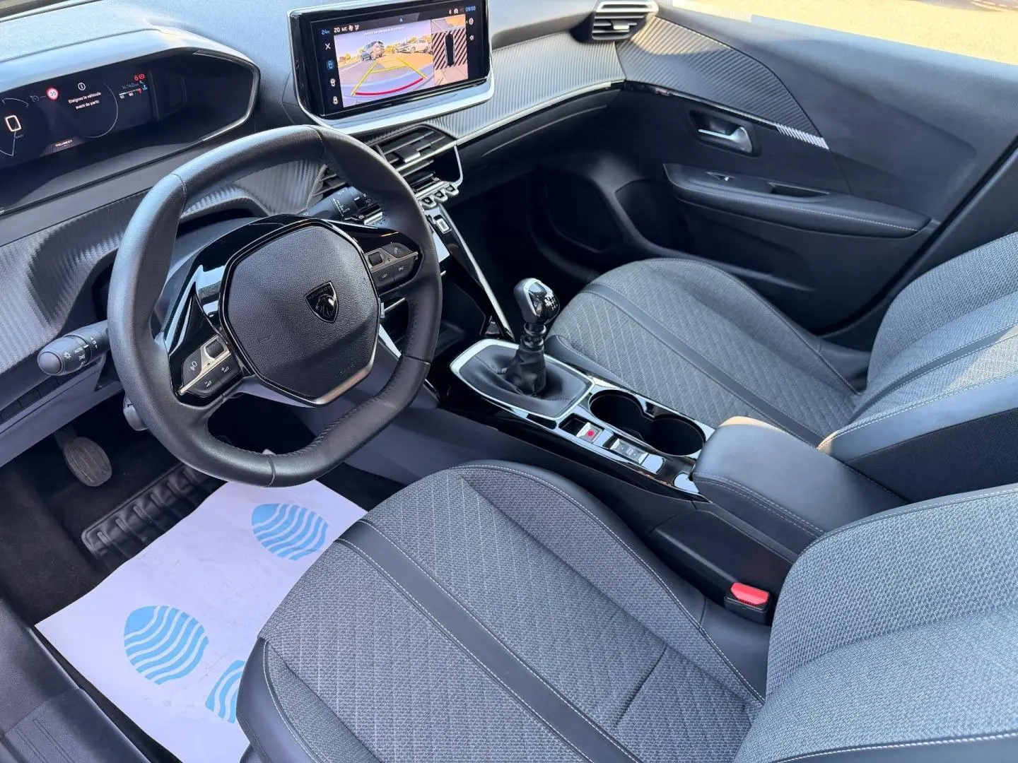 Vue intérieure côté conducteur de la Peugeot 208 1.2 essence 2025, volant compact et sièges tissu gris avec console centrale noire.
