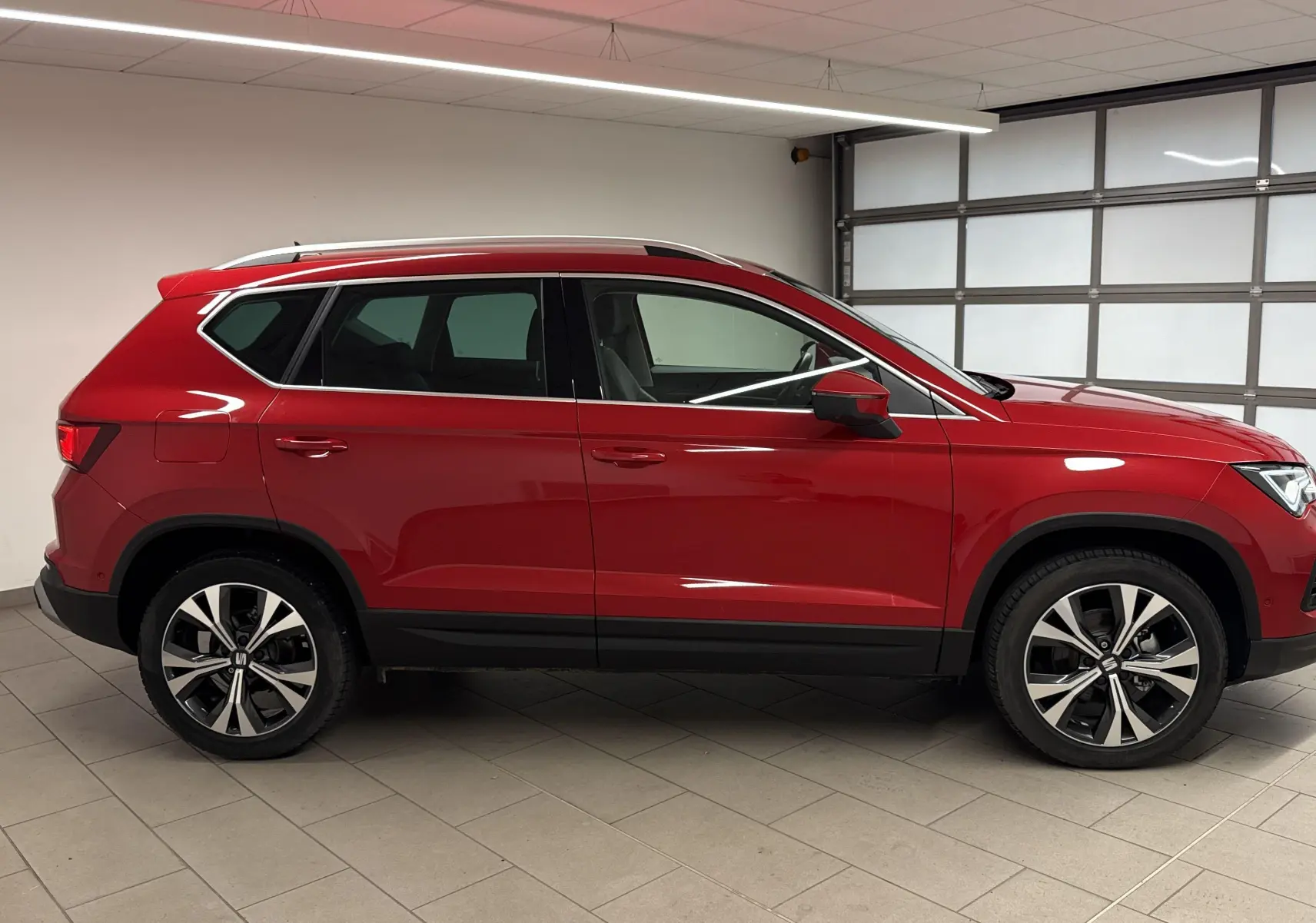 Vue latérale droite d'un SUV SEAT Ateca rouge velvet 2024 avec jantes alliage et toit ouvrant en intérieur garage.