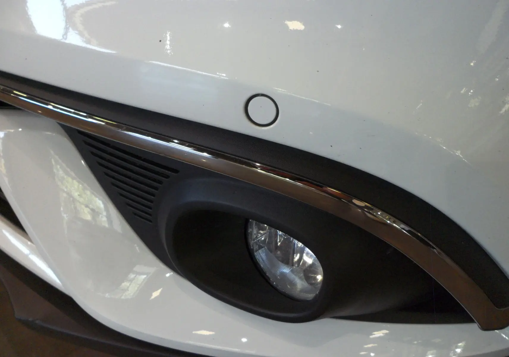 Gros plan sur le feu antibrouillard avant droit blanc glacier du Renault Grand Scenic IV avec capteur de stationnement.