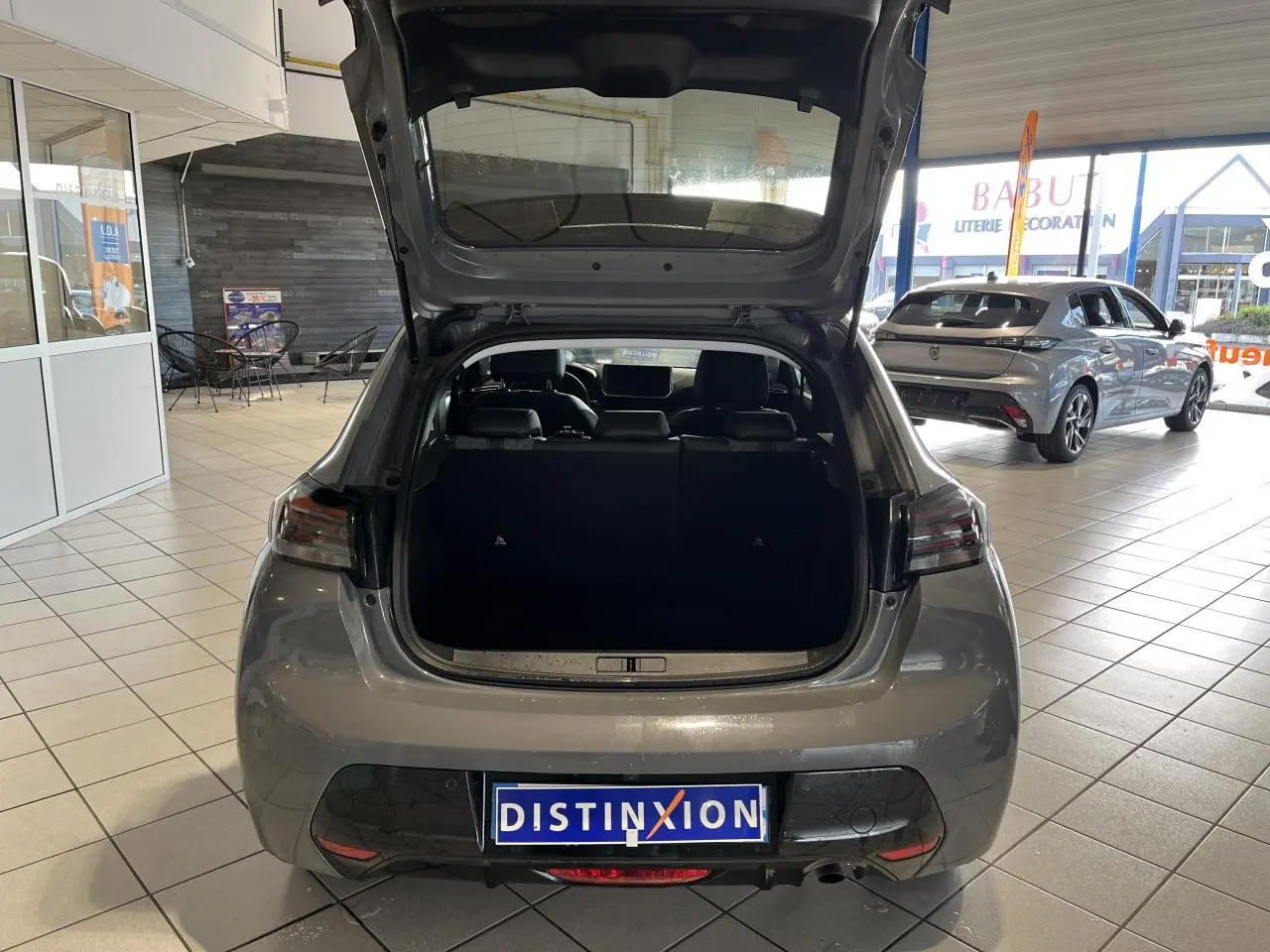 Vue arrière avec coffre ouvert d'une Peugeot 208 1.2 essence gris clair Selenium en showroom.