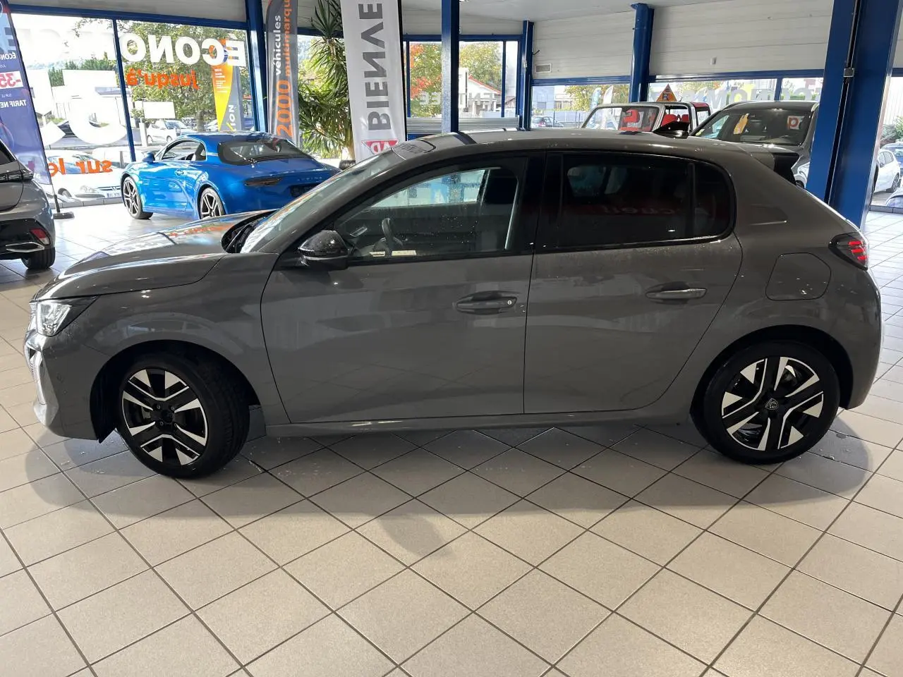 Profil côté gauche de la Peugeot 208 1.2 essence gris clair Selenium avec jantes alliage diamantées en showroom.