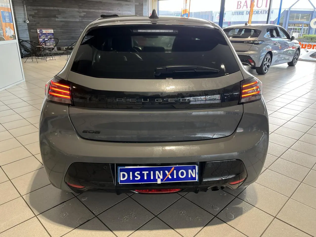 Vue arrière d'une Peugeot 208 gris clair Selenium avec feux arrière à LED et bandeau noir brillant en concession.