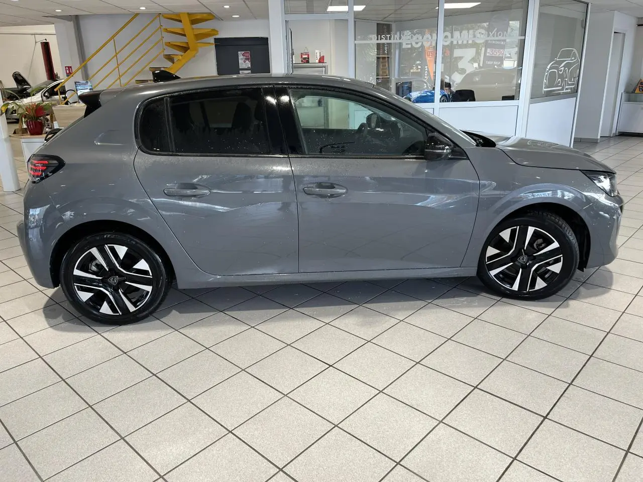 Peugeot 208 gris clair Selenium vue de profil côté gauche en intérieur avec jantes alliage bi-tons et vitres teintées.