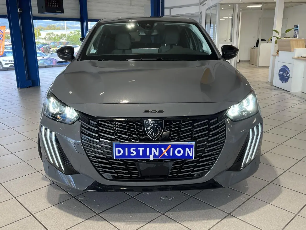 Vue avant d'une Peugeot 208 gris clair Selenium avec calandre noire et feux LED allumés en showroom.