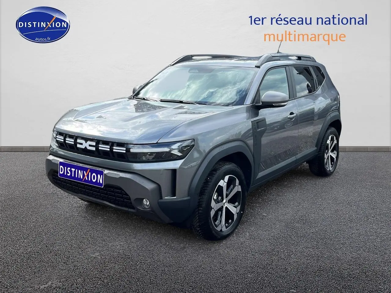 Dacia Duster gris schiste en 3/4 avant droit, avec jantes alu et barres de toit visibles.