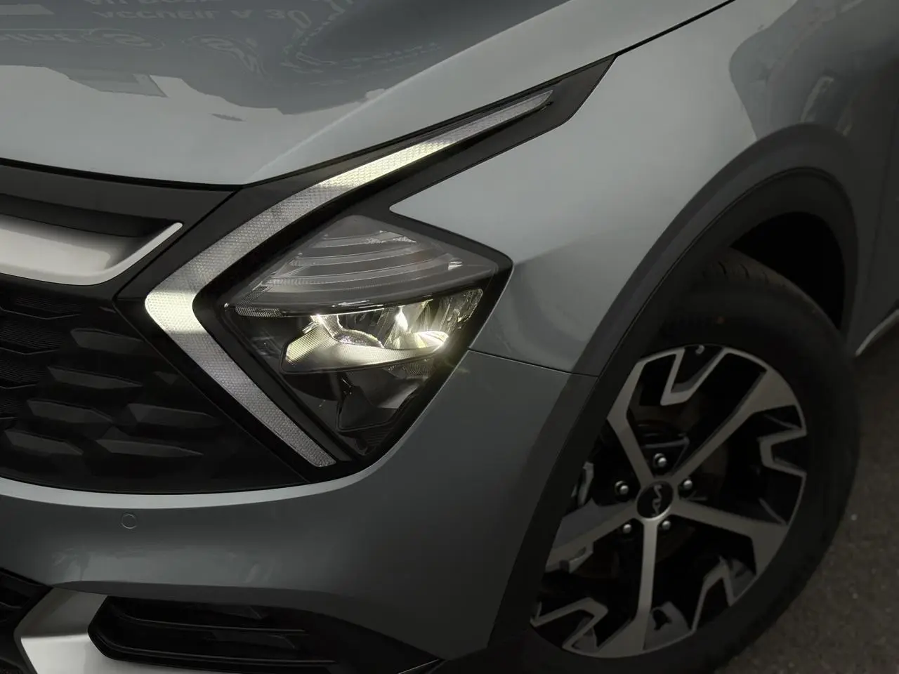 Gros plan sur le phare avant droit allumé et la jante alliage 18'' du Kia Sportage gris perle 2025.