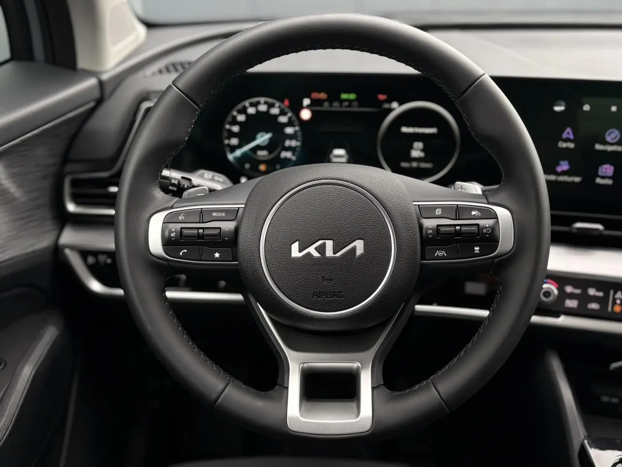 Volant cuir noir du Kia Sportage 2025 avec commandes intégrées, tableau de bord numérique et écran tactile visible en arrière-plan.