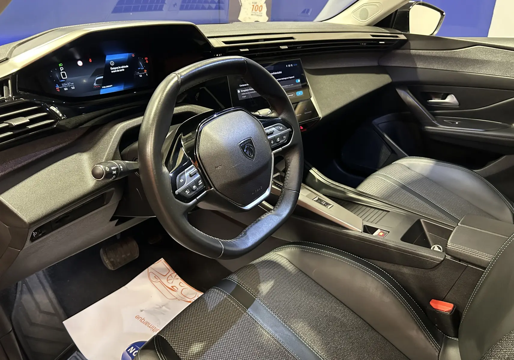 Vue intérieure côté conducteur de la Peugeot 308 2024 avec volant cuir, tableau de bord numérique et sièges noirs.