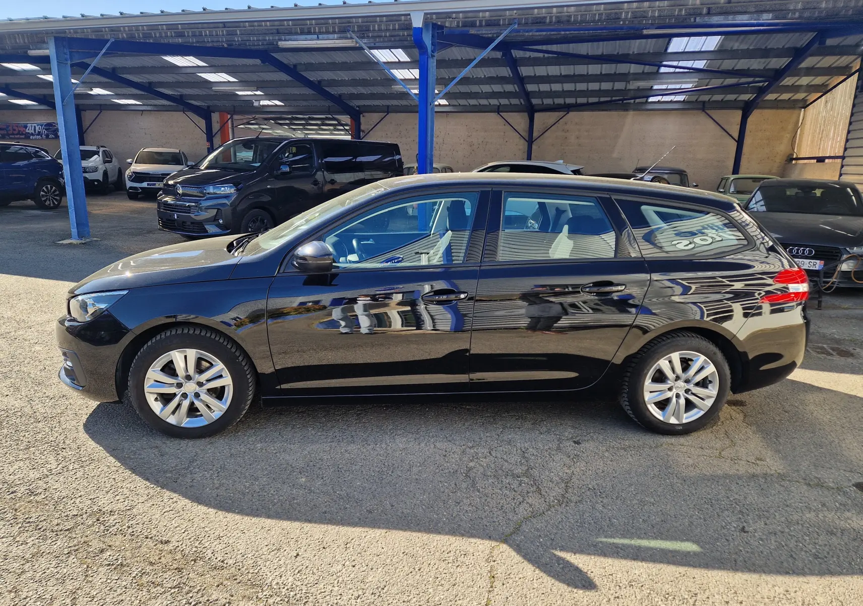 Peugeot 308 SW noire vue de profil côté gauche, stationnée en extérieur sous abri avec jantes alliage 16 pouces.