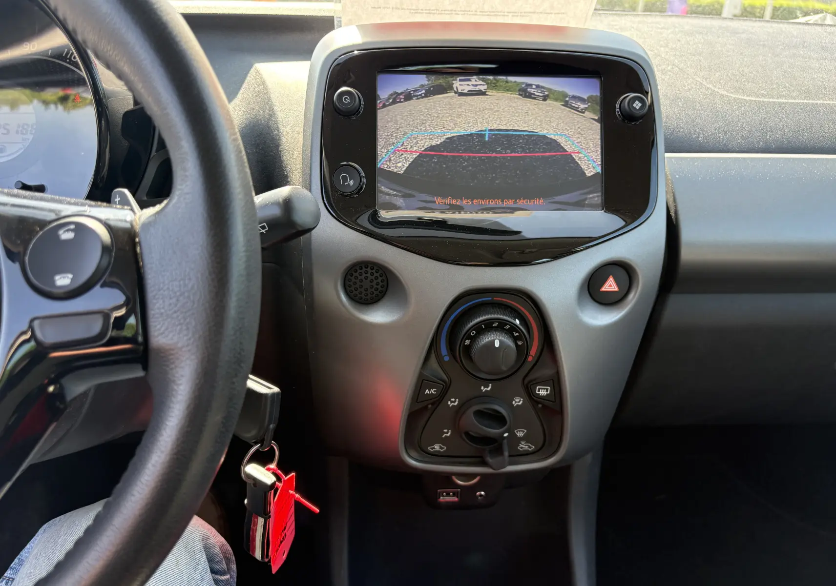 Vue intérieure centrée sur la console centrale d'une Toyota Aygo MC blanche, écran de caméra de recul et commandes climatisation visibles.