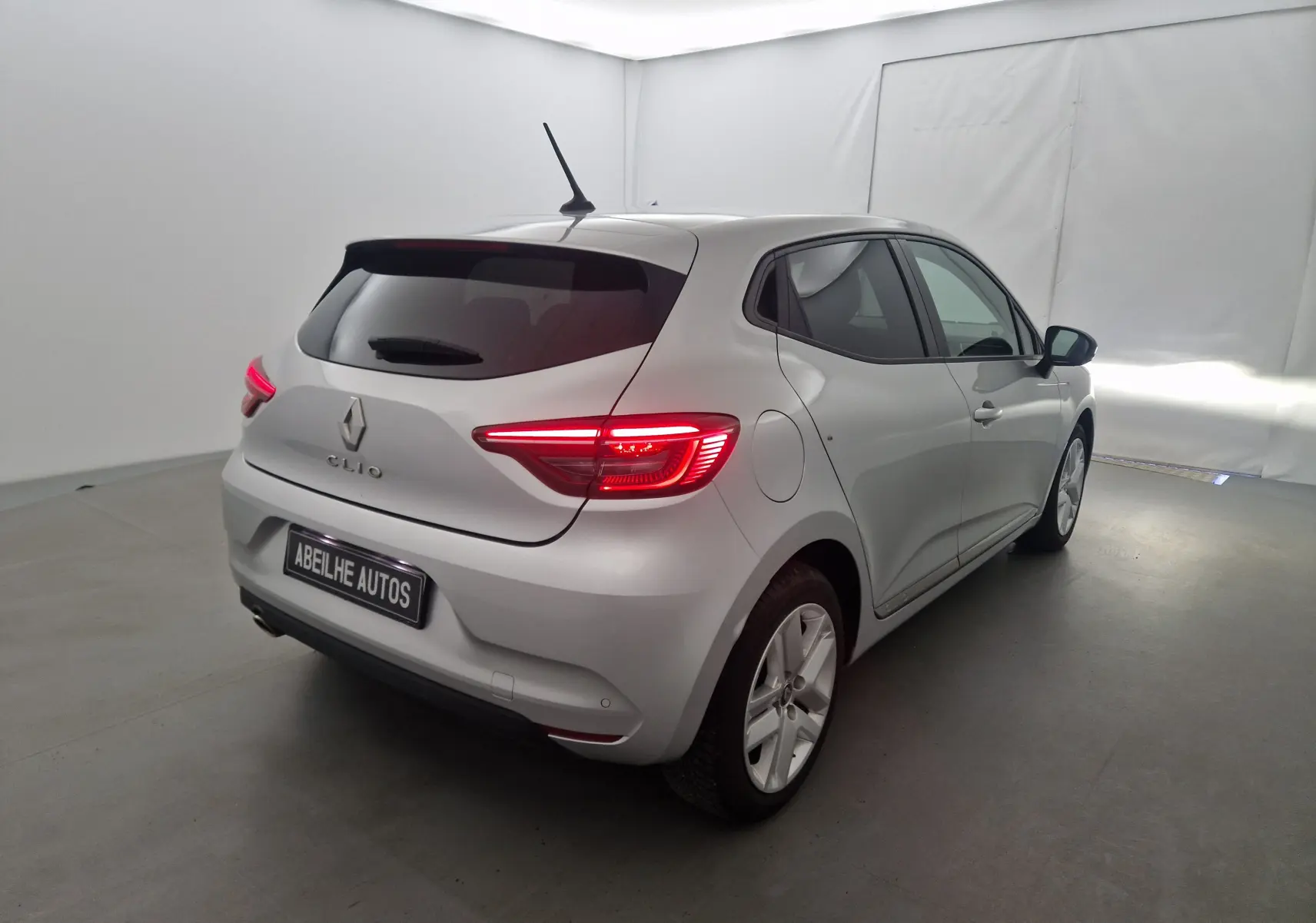 Vue 3/4 arrière droite d'une Renault Clio V gris platine avec feux arrière allumés dans un studio blanc.