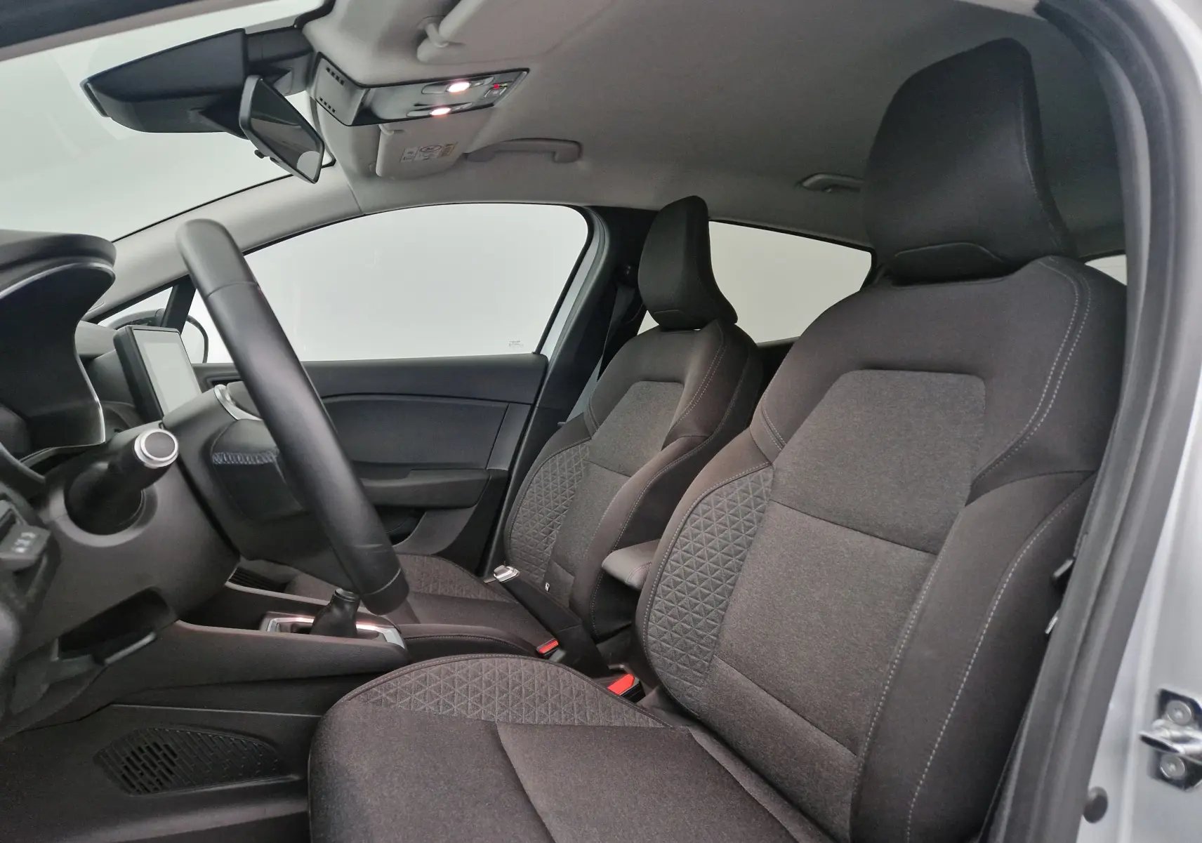 Vue intérieure côté conducteur de la Renault Clio V gris platine, sièges tissu noir avec motifs géométriques et volant visible.