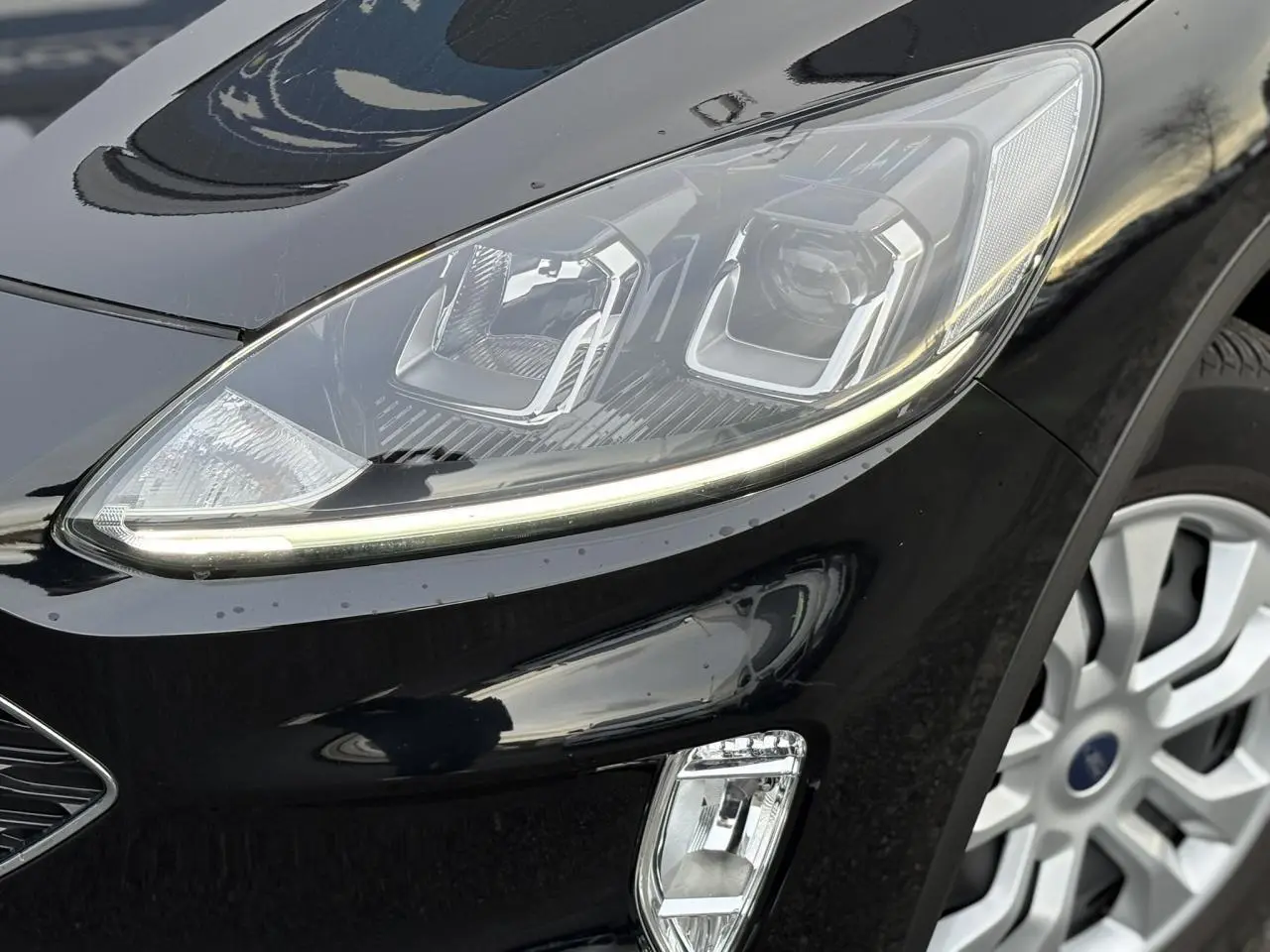 Gros plan sur le phare avant droit du Ford Kuga noir Obsidien 2024 avec jante alliage visible.