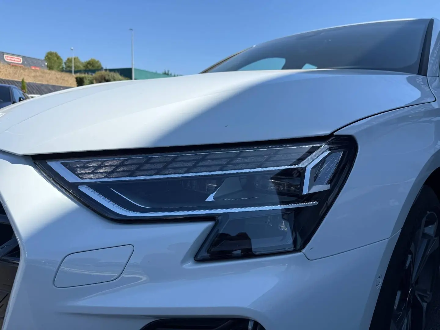 Gros plan sur le phare avant droit de l'Audi A3 Sportback blanc Glacier métallisé, avec jante alu visible en arrière-plan.