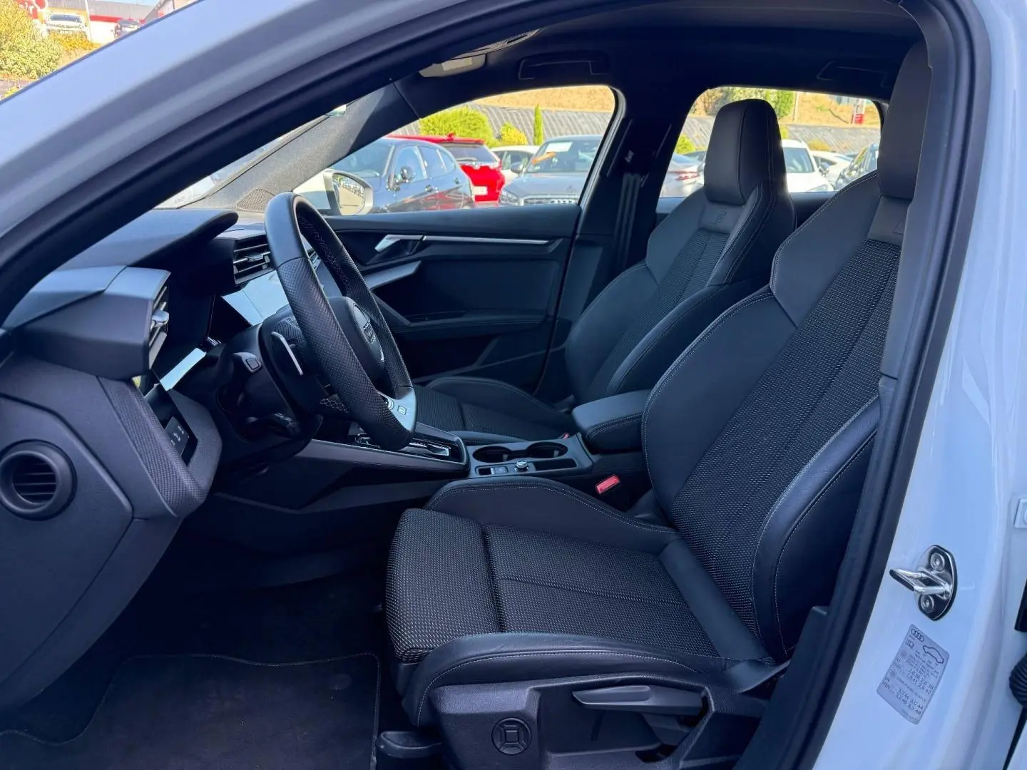 Vue intérieure côté conducteur de l'Audi A3 Sportback 2025 blanche, sièges sport noirs et volant multifonction cuir.