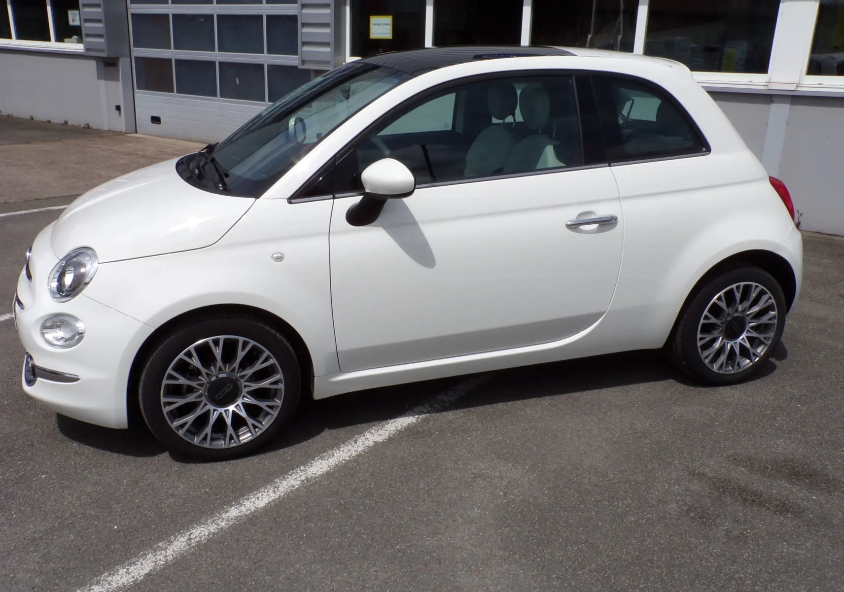 Profil droit d'une Fiat 500 blanche 2019 avec jantes alliage bicolores et rétroviseurs blancs stationnée sur un parking.