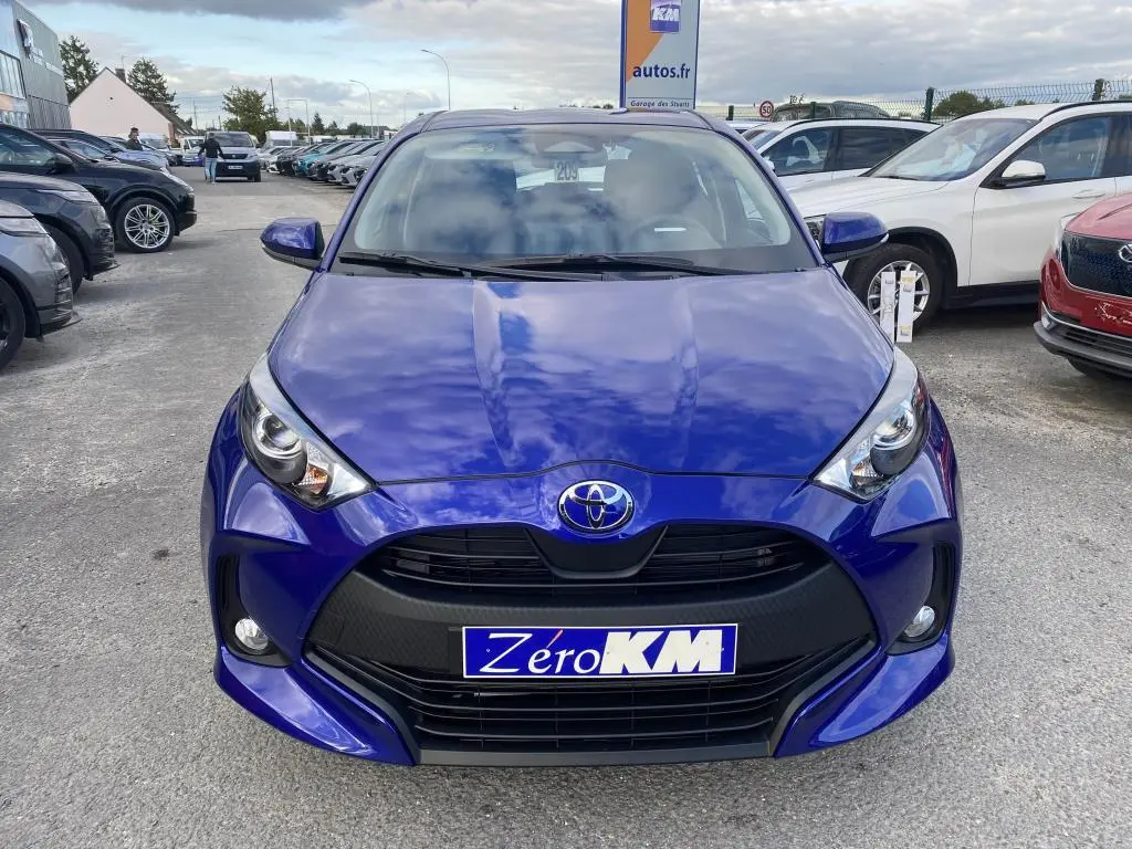 Vue frontale d'une Toyota Yaris 2025 bleu Genièvre avec calandre noire et phares LED allumés sur parking.