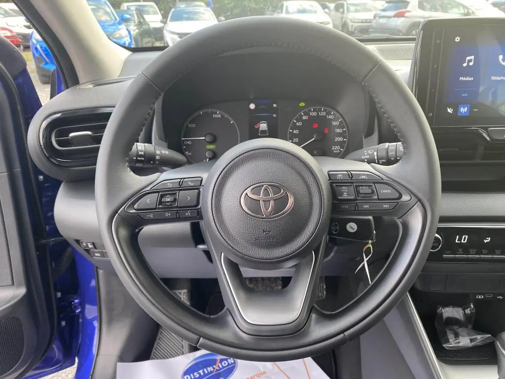 Vue centrée sur le volant noir de la Toyota Yaris 2025, tableau de bord et écran tactile avec portière bleu Genièvre ouverte.