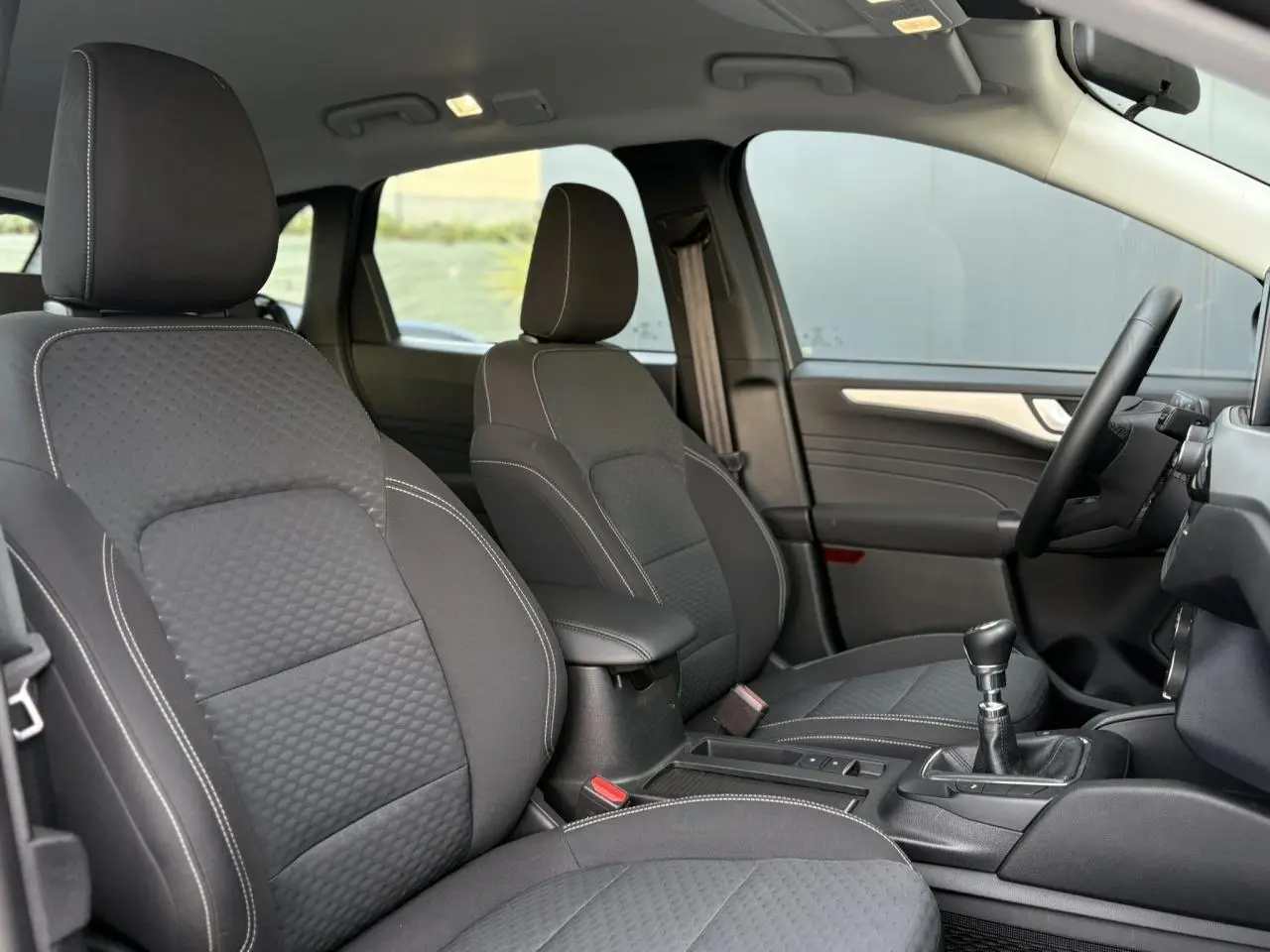 Vue intérieure côté droit du Ford Kuga 2024 noir Obsidien, montrant les sièges avant en tissu avec surpiqûres blanches et la boîte manuelle.