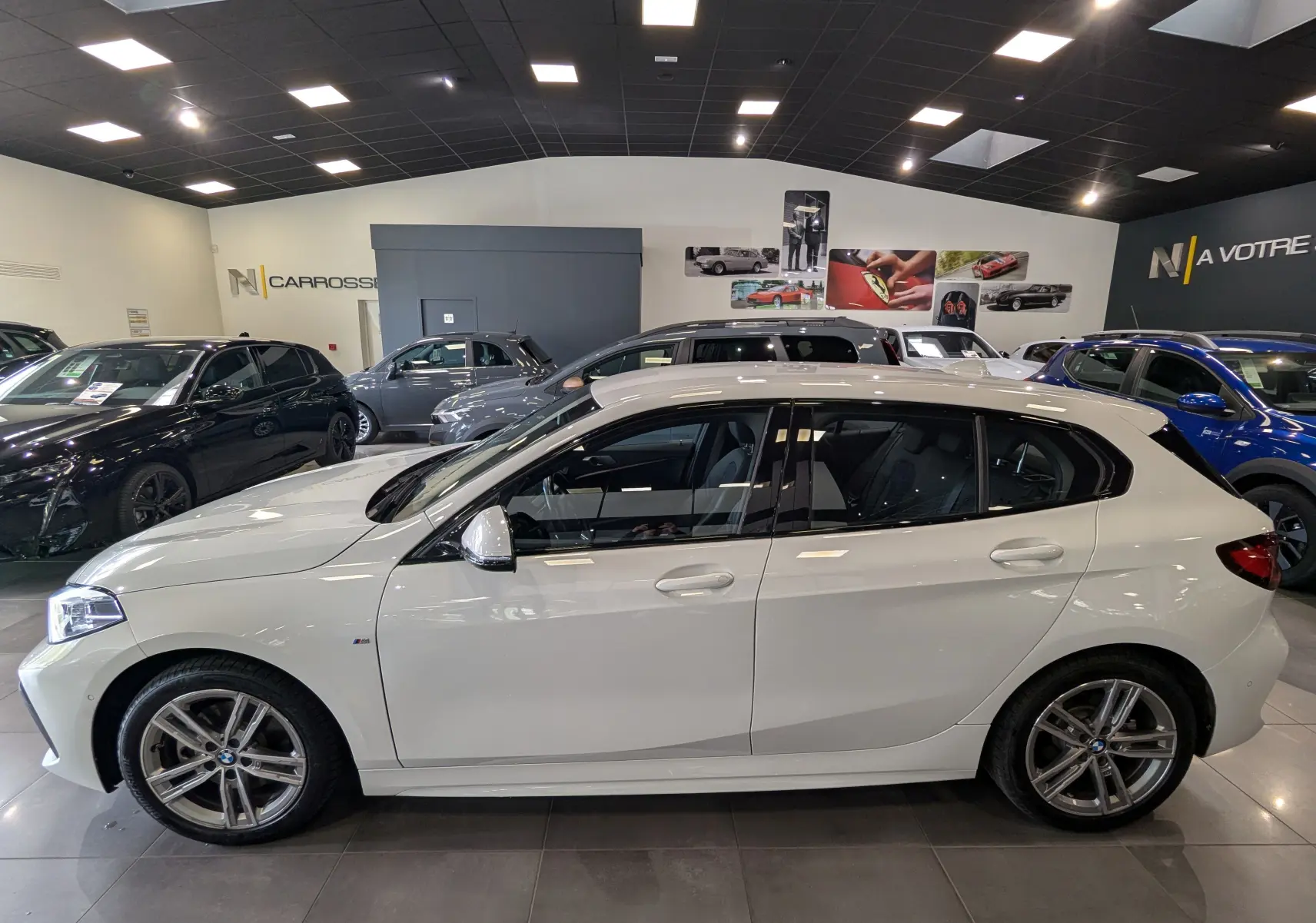 BMW Série 1 F40 18d blanc alpin vue de profil côté gauche en intérieur avec jantes M Sport et rétroviseurs blancs.