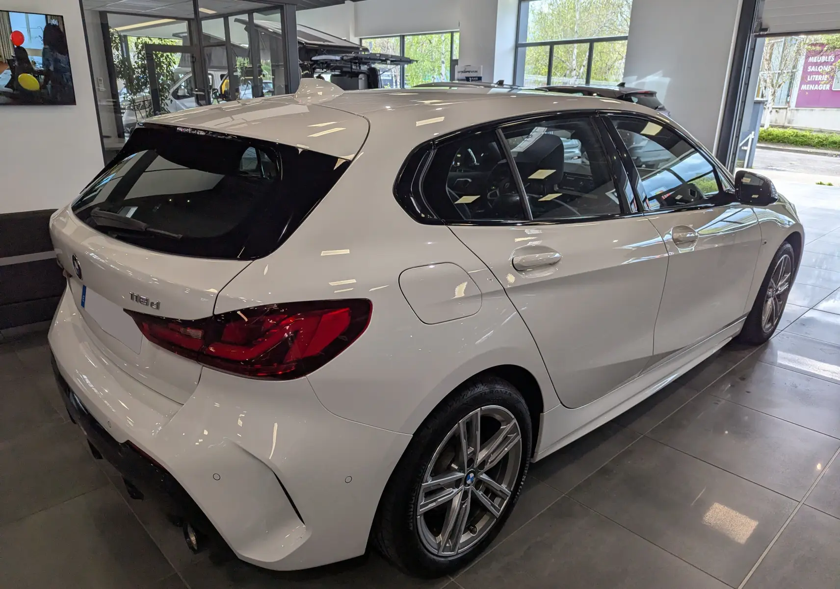 BMW Série 1 F40 18d blanc alpin vue 3/4 arrière droit en intérieur avec jantes alliage et feux LED distinctifs.