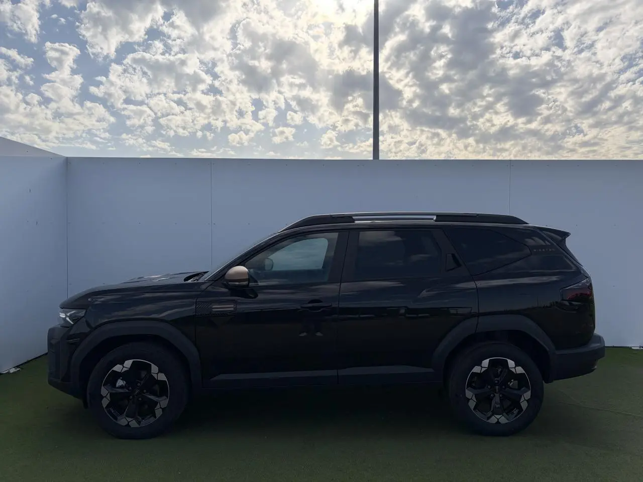 Profil côté gauche du Dacia Bigster noir nacré 2026 avec jantes alliage et barres de toit visibles sous ciel nuageux.