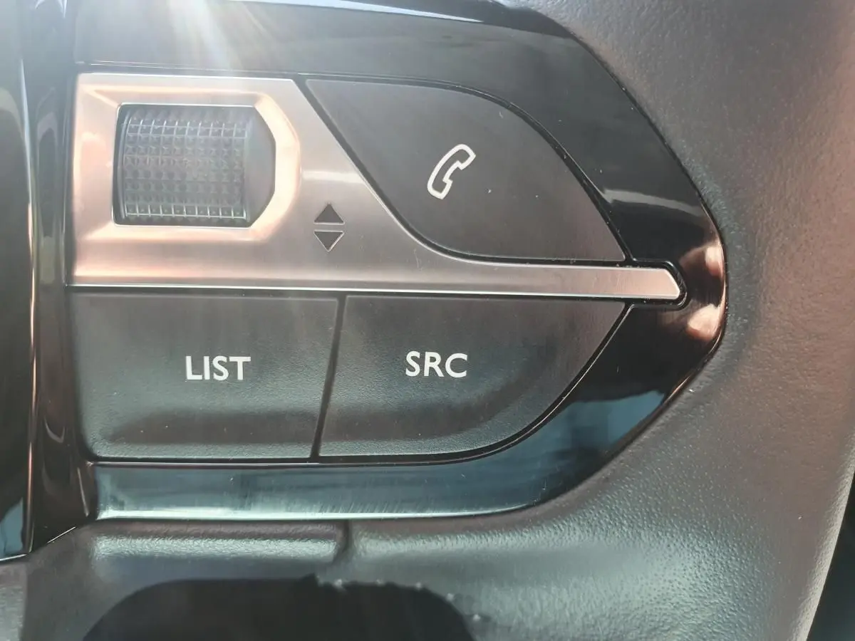 Gros plan sur les commandes au volant côté gauche de la Peugeot 208 gris Artense, boutons LIST, SRC et téléphone.