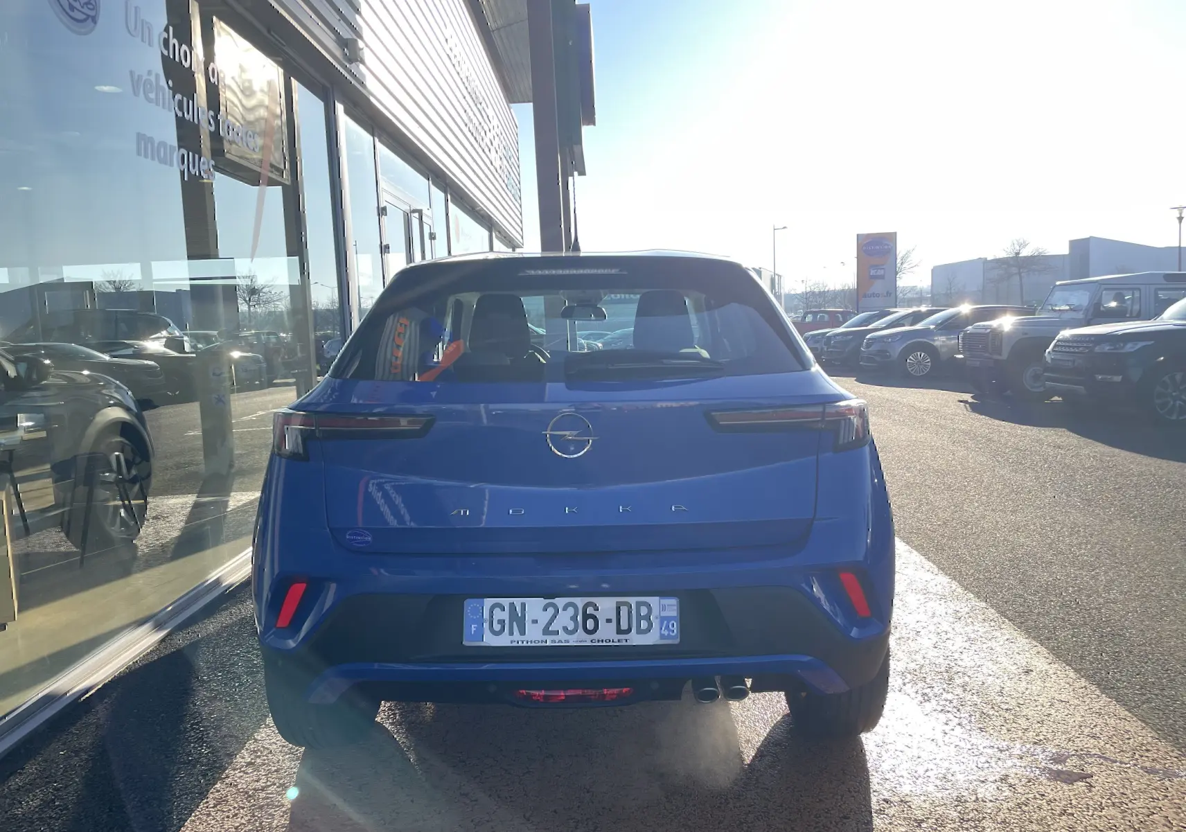 Vue arrière d'un Opel Mokka bleu Voltak 2023 garé devant une vitrine, avec feux arrière LED allumés.