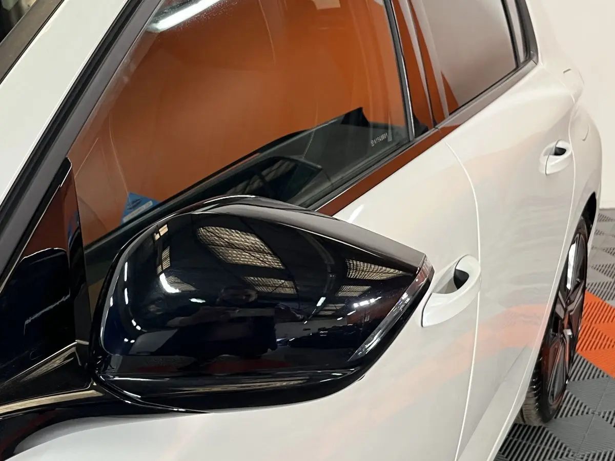 Gros plan sur le rétroviseur noir brillant côté gauche d'une Peugeot 308 GT PHEV blanc nacré, avec porte et vitre arrière visibles.