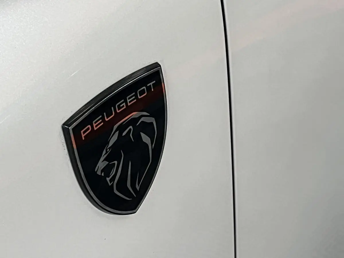 Gros plan sur l'écusson Peugeot noir et rouge apposé sur la carrosserie blanc nacré d'une 308 GT PHEV 2022.