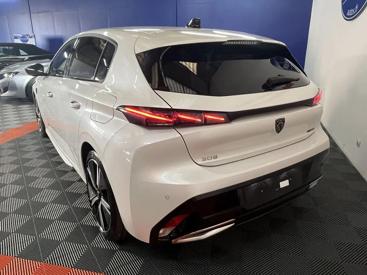 Vue 3/4 arrière droite d'une Peugeot 308 GT PHEV 180 blanche nacrée avec feux arrière LED allumés en intérieur showroom.