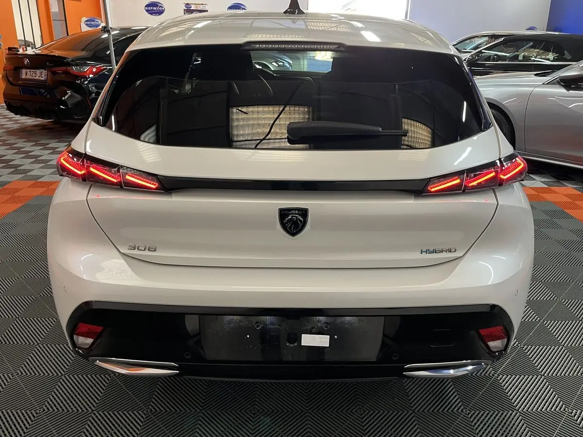 Vue arrière d'une Peugeot 308 GT PHEV blanche nacrée avec feux LED allumés et logo noir au centre du hayon.