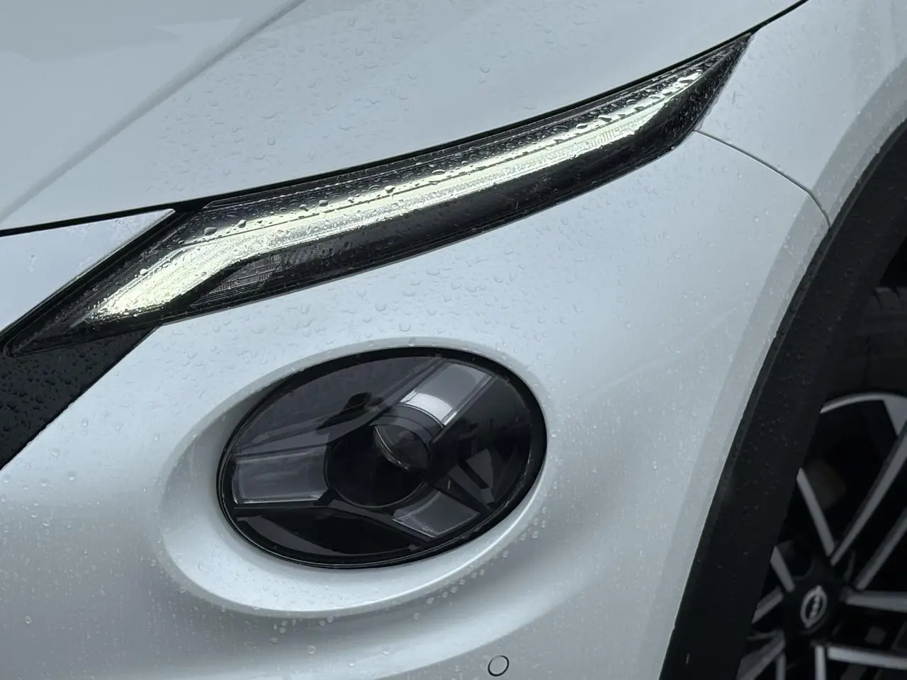 Gros plan sur le phare avant droit et la jante alliage 17'' d'un Nissan Juke blanc nacré sous la pluie.