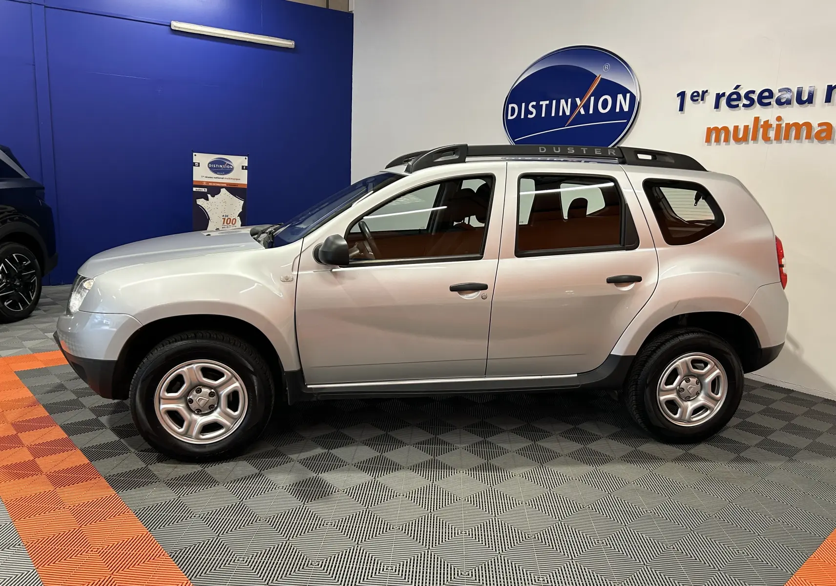 Profil latéral droit d'un Dacia Duster gris 2017, avec barres de toit et jantes acier, en intérieur showroom.
