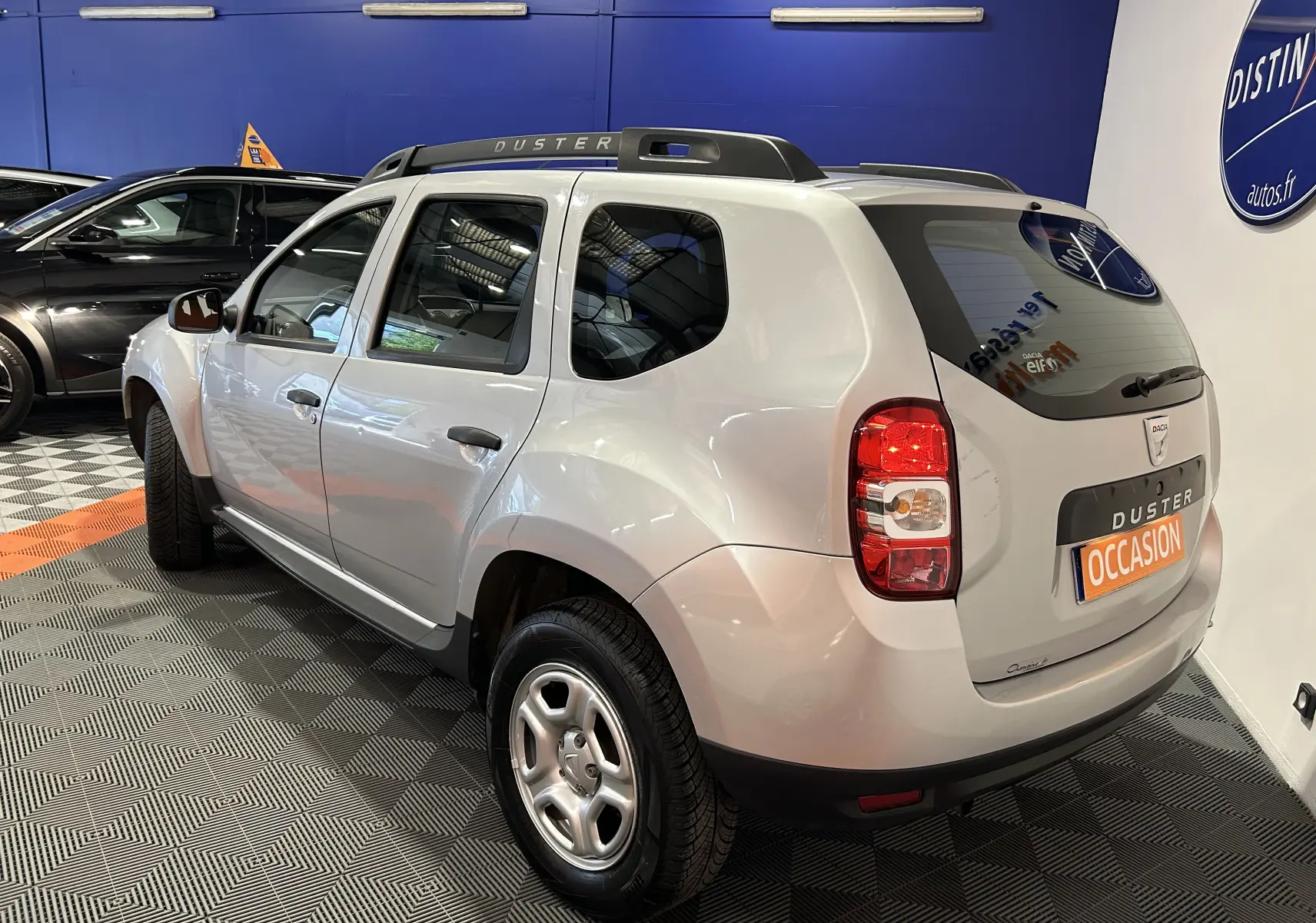 Vue 3/4 arrière droite d'un Dacia Duster gris 2017 avec barres de toit et plaque "OCCASION" en concession.